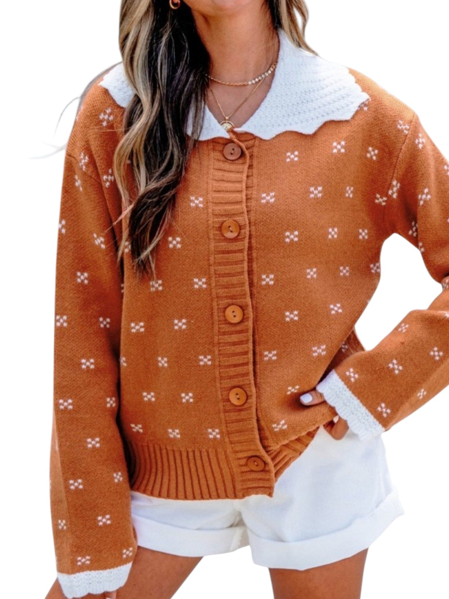 A woman wearing a rust-orange knit cardigan with a white scalloped collar, white cuffs, and small white cross motifs, paired with white shorts.