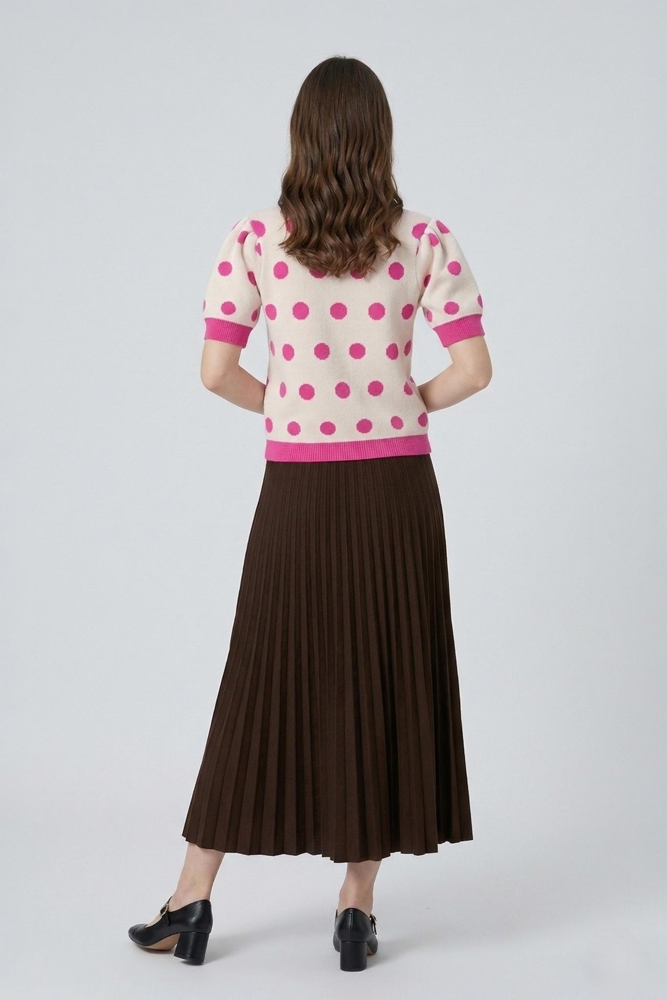 Back view of a model wearing a pink polka dot knit top with puff sleeves, paired with a dark brown pleated skirt.
