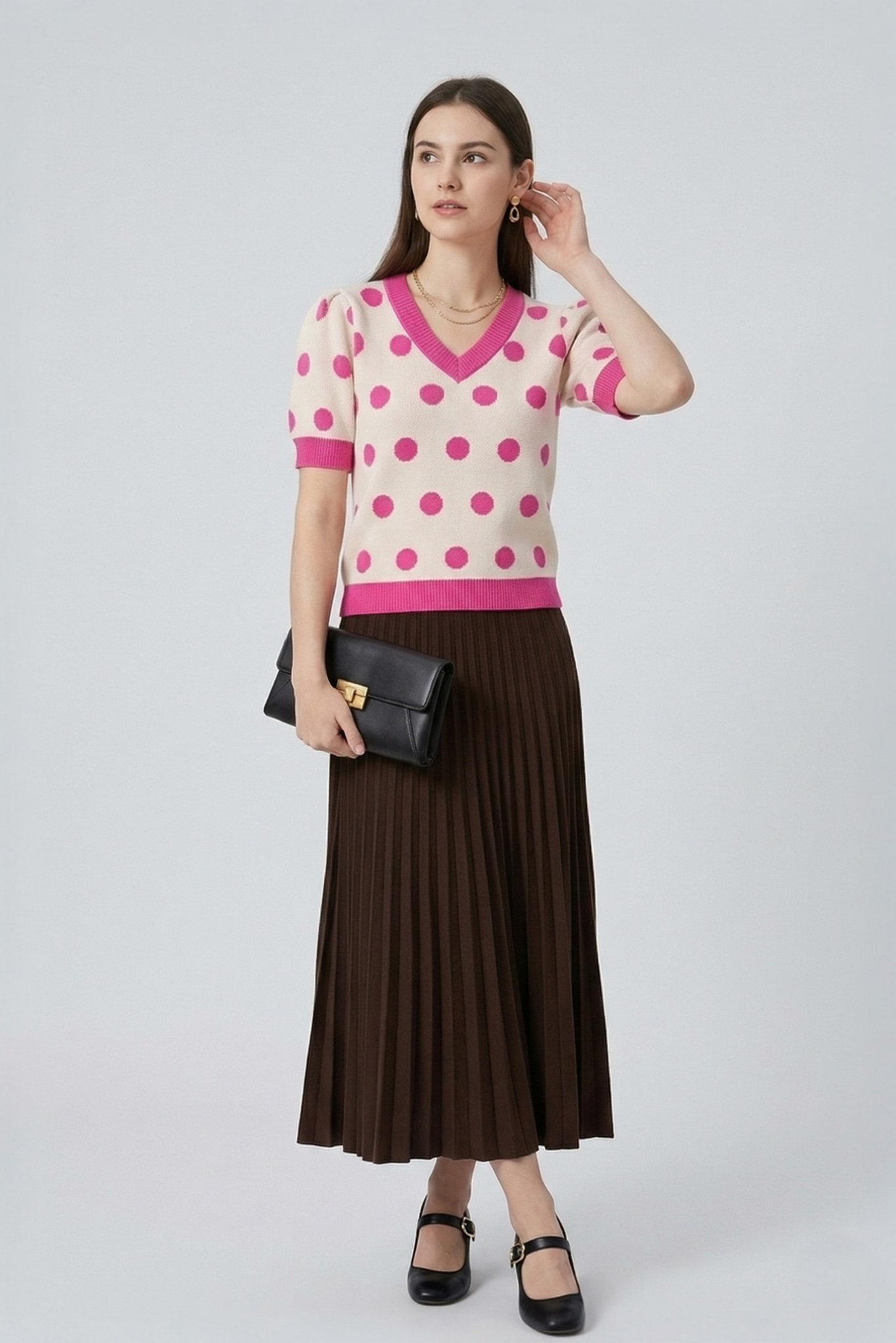 Model holding a black leather clutch while wearing a cream and pink polka dot knit top and brown pleated midi skirt.