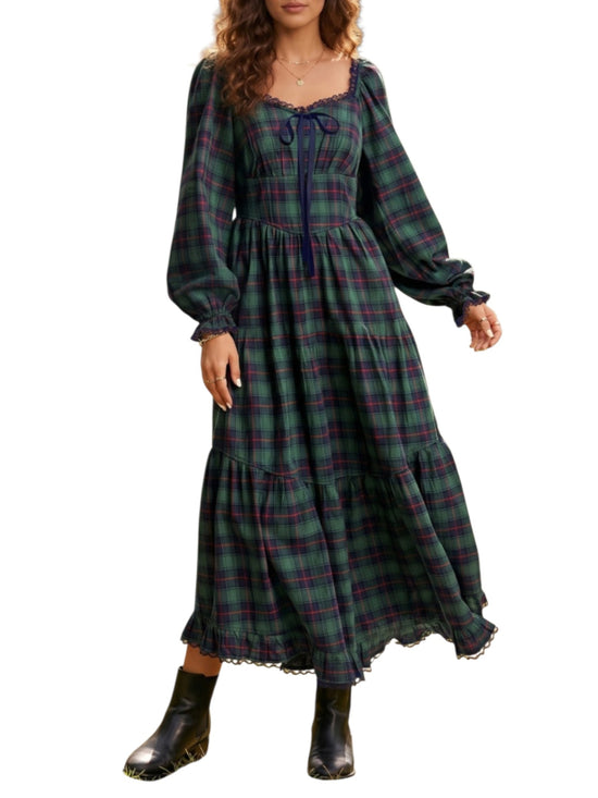 A woman wearing a long-sleeve plaid maxi dress with a tie neckline and tiered skirt, paired with black boots, against a plain white background.