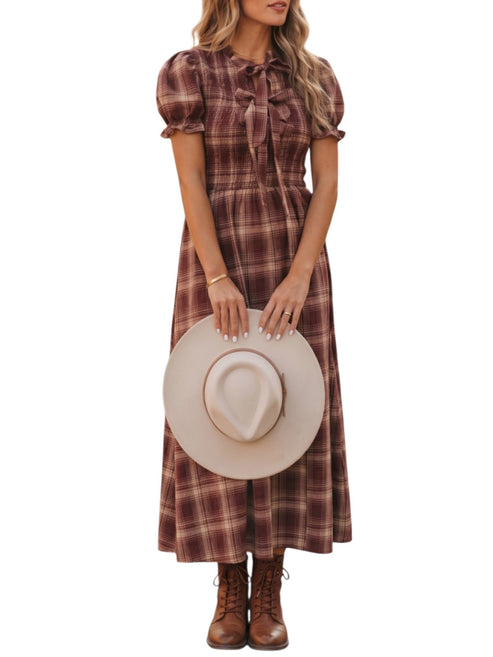 A woman wearing a brown and burgundy plaid midi dress with puff sleeves and a bow at the neckline, holding a beige wide-brim hat and wearing brown lace-up boots against a white background.