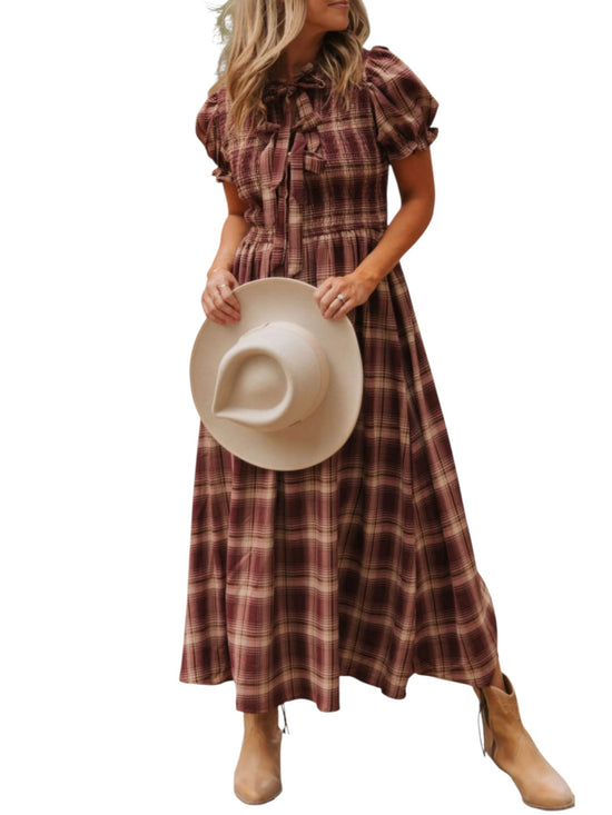 A woman wearing a long plaid midi dress in brown and burgundy tones, holding a beige wide-brim hat, against a plain white background.
