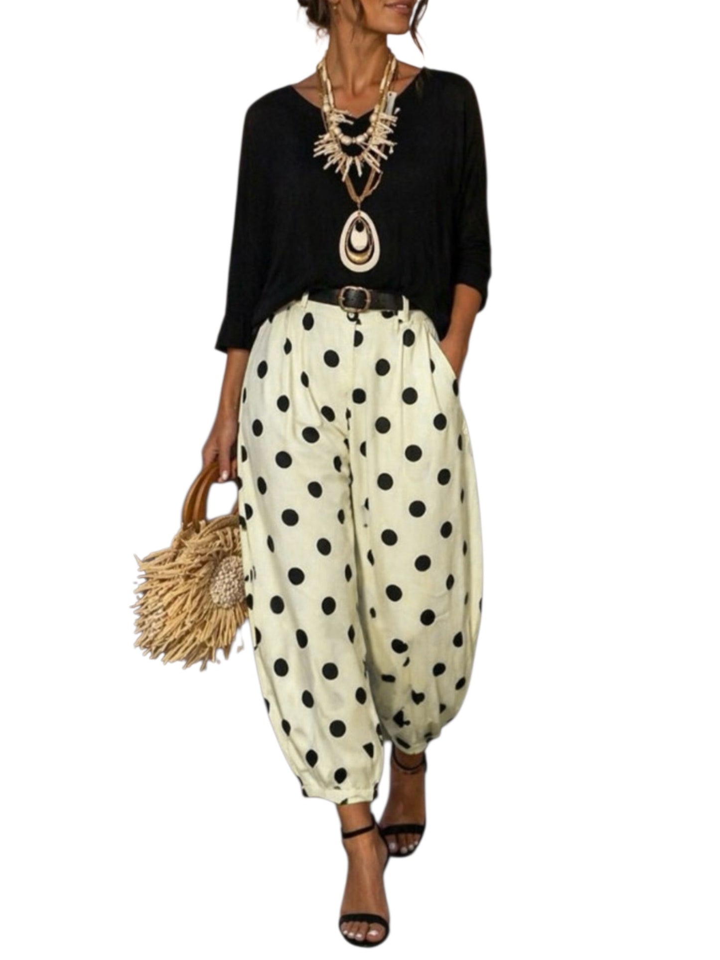 A woman modeling loose, balloon-style pants with large black polka dots on an off-white base, paired with a black top, statement necklace, and straw handbag against a plain white background.