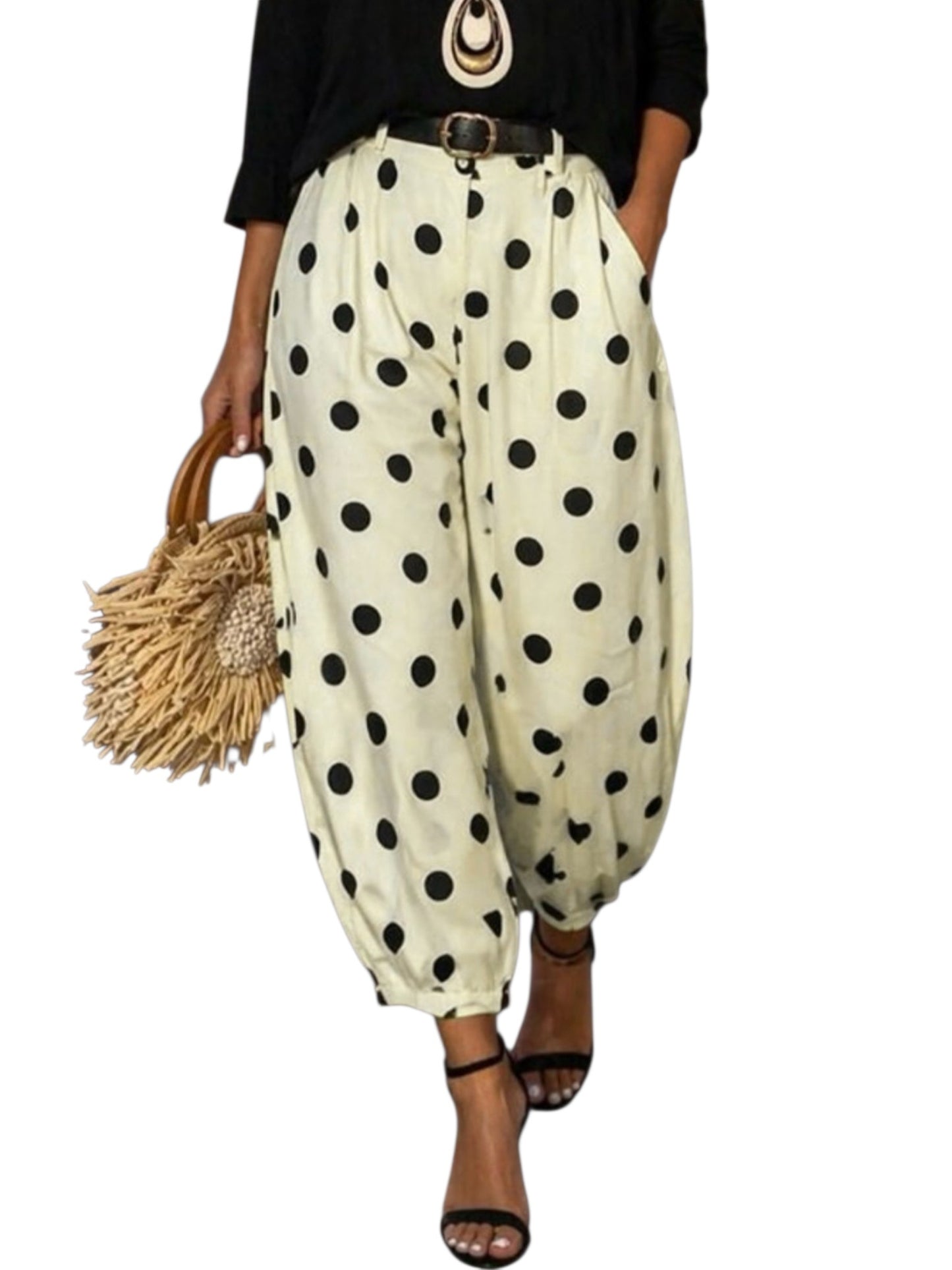 A person wearing loose, balloon-style pants with large black polka dots on a cream background, paired with a black top, brown belt, and black heeled sandals, while holding a woven straw handbag against a plain white background.
