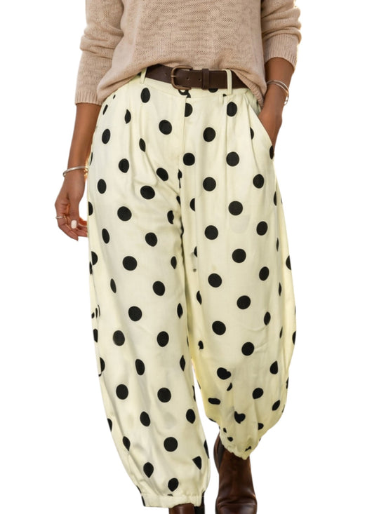 A person wearing loose, balloon-style pants with large black polka dots on a warm off-white background, paired with a beige sweater and brown belt, shown against a plain white studio background.