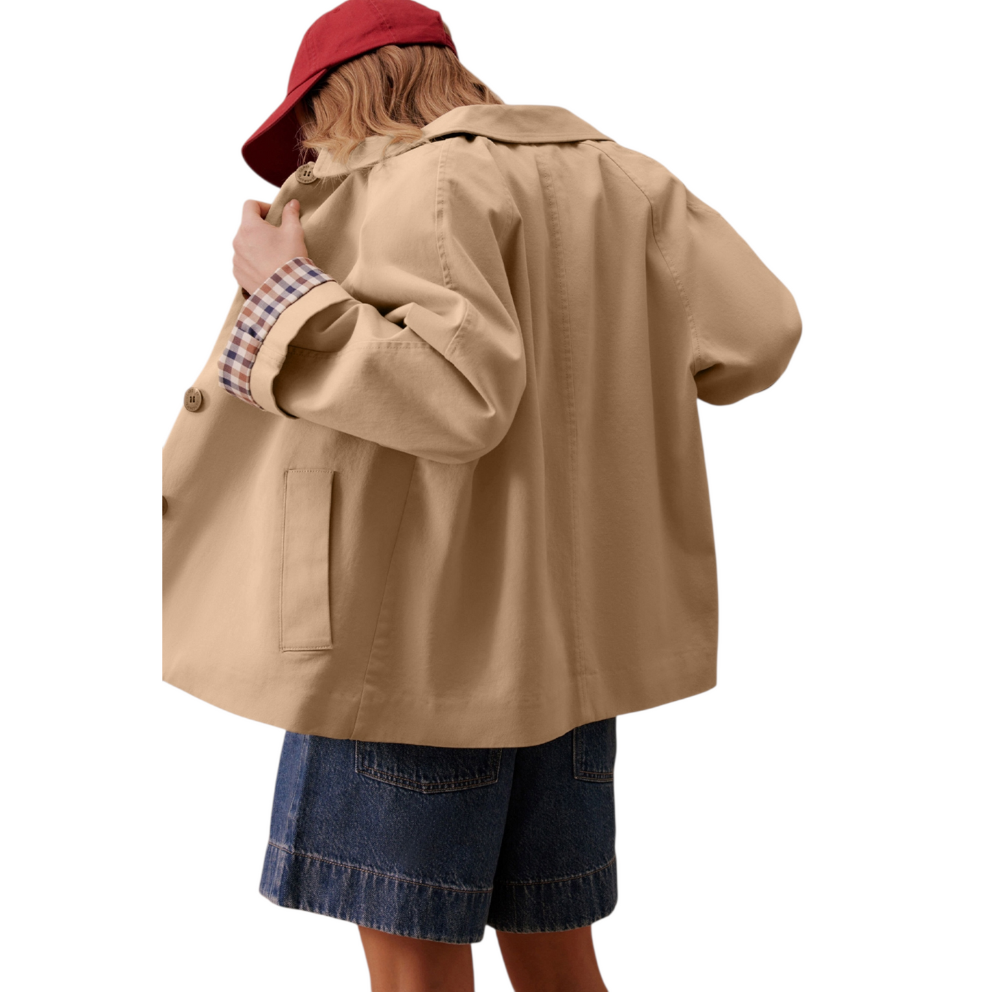 A woman wears a beige, short trench coat with the plaid lining visible on the turned-up cuffs, over a striped shirt and dark-wash denim shorts.