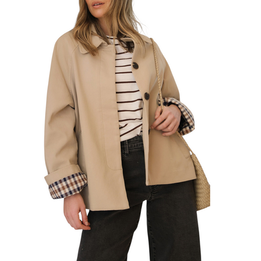 A woman wears a beige, short trench coat with the plaid lining visible on the turned-up cuffs, over a striped shirt and dark-wash jeans.