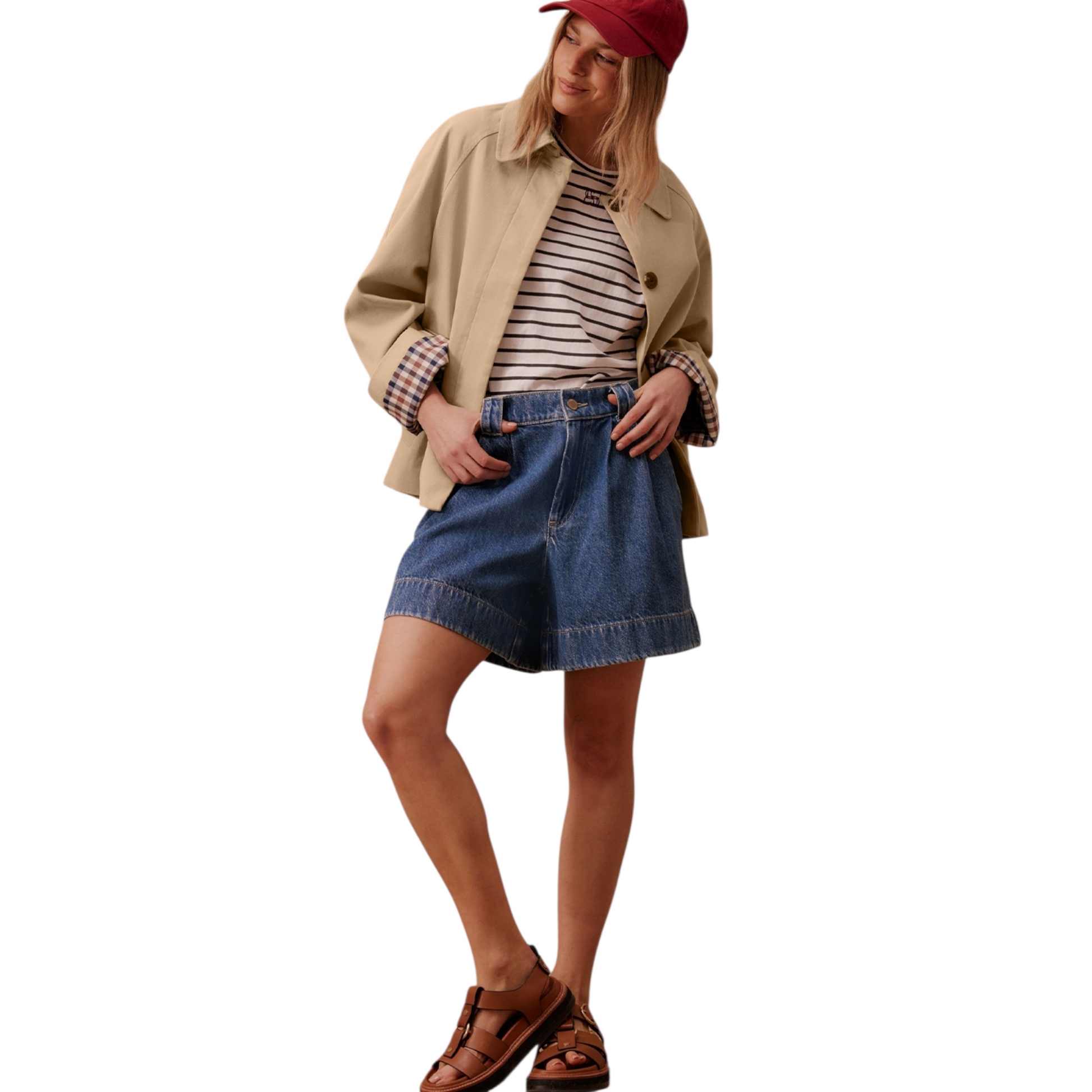 A woman wears a beige, short trench coat with the plaid lining visible on the turned-up cuffs, over a striped shirt and dark-wash denim shorts.
