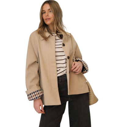 A woman wears a beige, short trench coat with the plaid lining visible on the turned-up cuffs, over a striped shirt and dark-wash jeans.