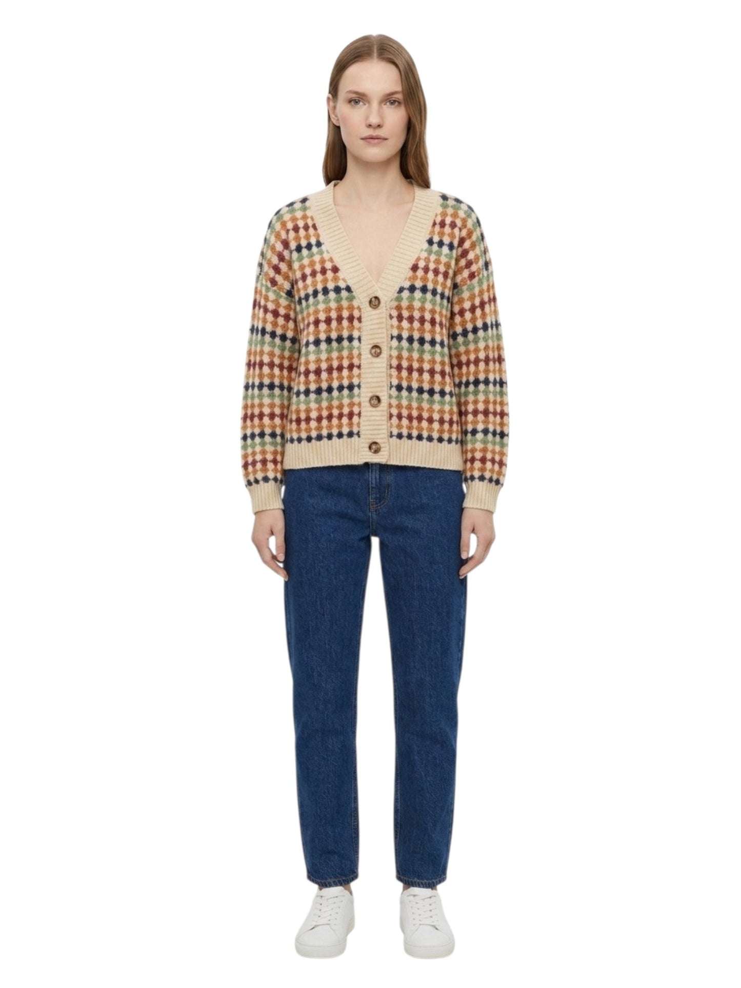 A model stands against a white background wearing a colorful knit cardigan with a beige base and a multicolor plaid pattern, paired with blue jeans and white sneakers.