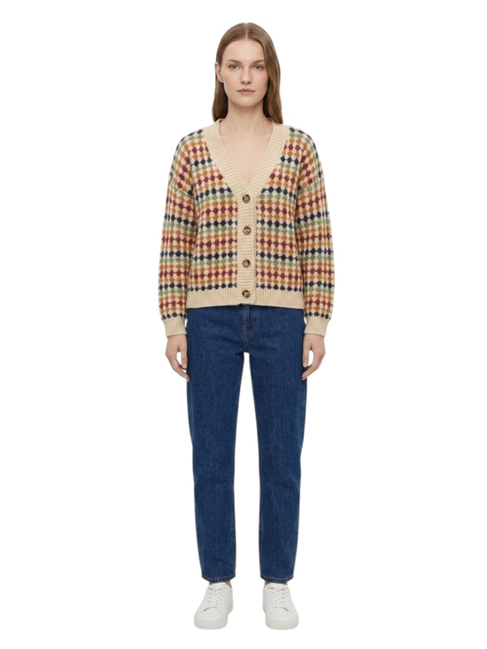 A model stands against a white background wearing a colorful knit cardigan with a beige base and a multicolor plaid pattern, paired with blue jeans and white sneakers.