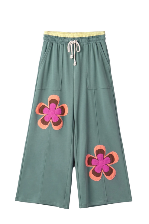 A professional flat lay front view of the orange wide-leg lounge pants, showcasing the oversized teal, pink, and blue floral patches and the yellow contrast waistband.