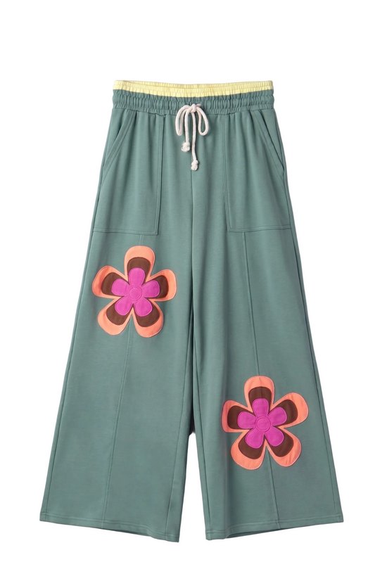 A professional flat lay front view of the orange wide-leg lounge pants, showcasing the oversized teal, pink, and blue floral patches and the yellow contrast waistband.
