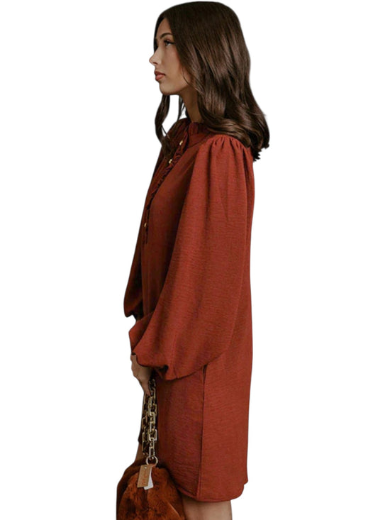 A woman shown in side profile wearing a rust-orange long-sleeve top with voluminous balloon sleeves and a high neckline, paired with a matching rust-orange skirt or dress, holding a brown handbag against a plain white background.