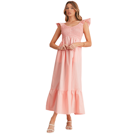 A woman wearing a peach-pink cotton maxi dress featuring a shirred elastic bodice, ruffle cap sleeves, and a tiered A-line skirt.