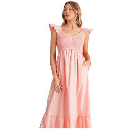 Model wearing a flowy peach cotton summer dress with ruffle sleeves and a smocked bodice, styled with white strappy heels. Half view.