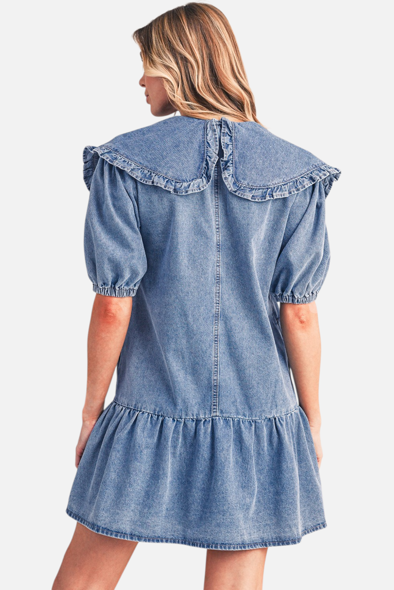 Rear view of a model wearing a blue denim mini dress, highlighting the large ruffled collar, relaxed drop-waist, and tiered ruffle hemline.