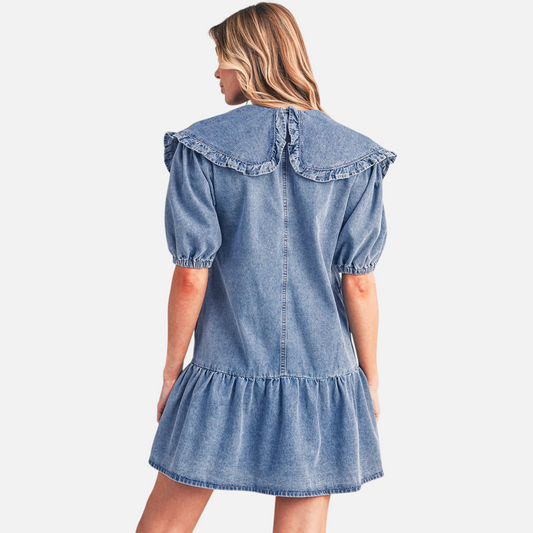 Rear view of a model wearing a blue denim mini dress, highlighting the large ruffled collar, relaxed drop-waist, and tiered ruffle hemline.