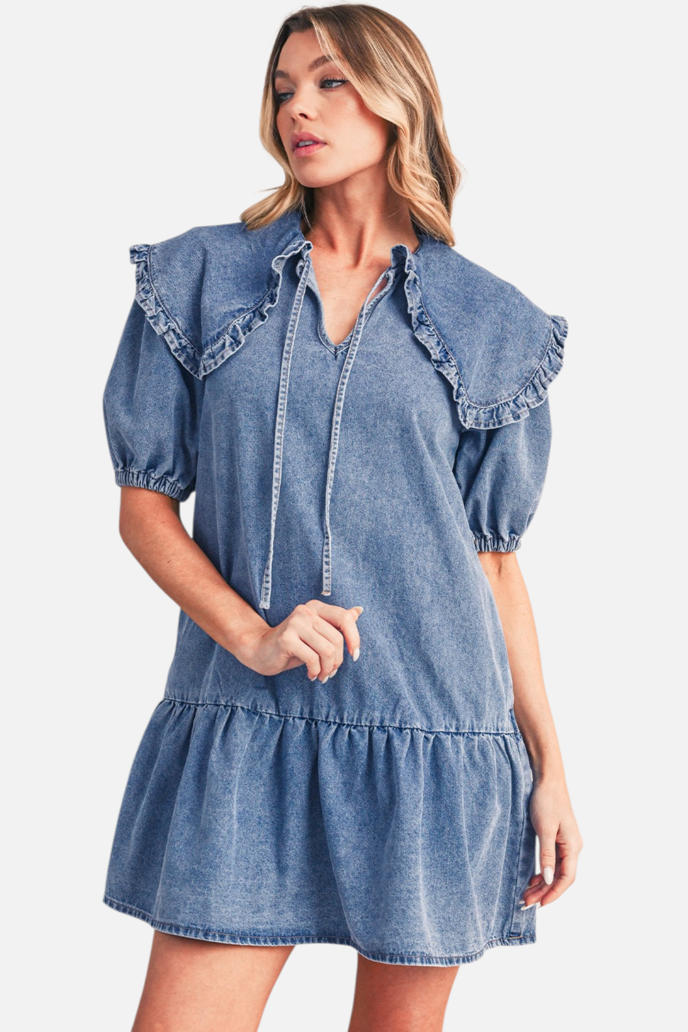 Front view of a model wearing a blue denim mini dress, highlighting the large ruffled collar, relaxed drop-waist, and tiered ruffle hemline.