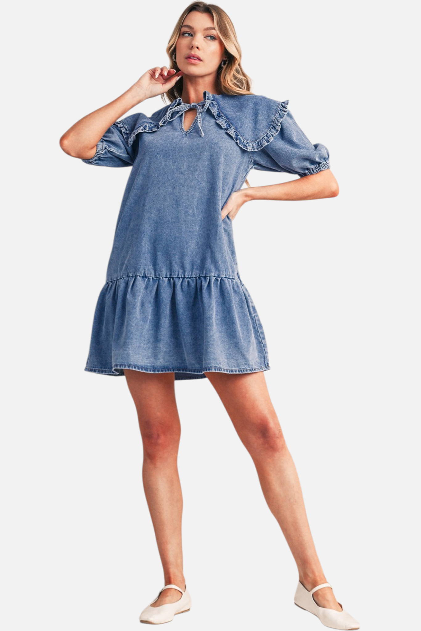 Full body view of a model wearing a blue denim mini dress, highlighting the large ruffled collar, relaxed drop-waist, and tiered ruffle hemline.