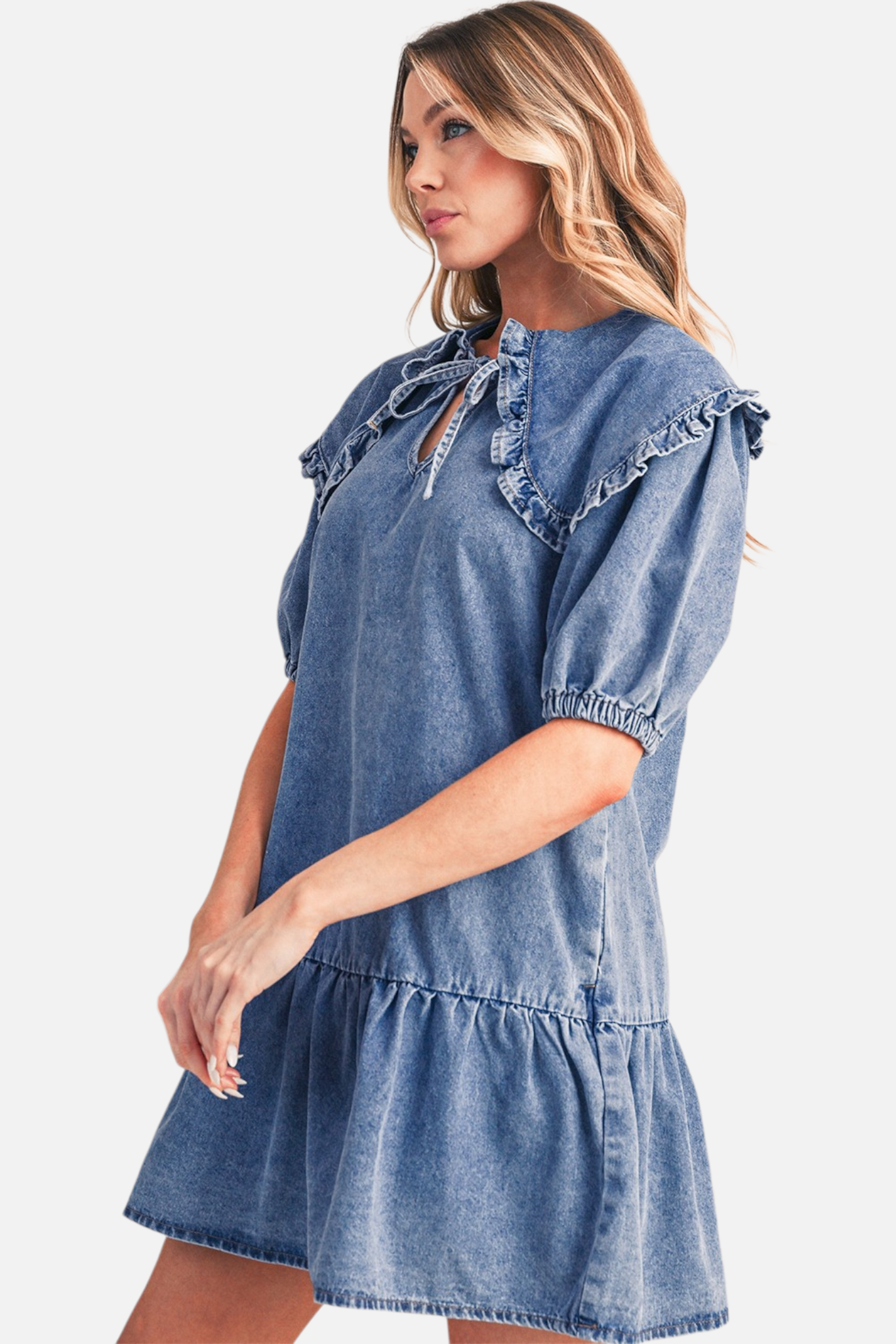 Side view of a model wearing a blue denim mini dress, highlighting the large ruffled collar, relaxed drop-waist, and tiered ruffle hemline.