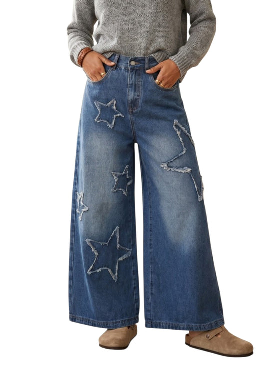 A person wearing blue wide-leg denim jeans with large embroidered star patches, paired with a gray sweater and tan shoes, shown against a plain white background.