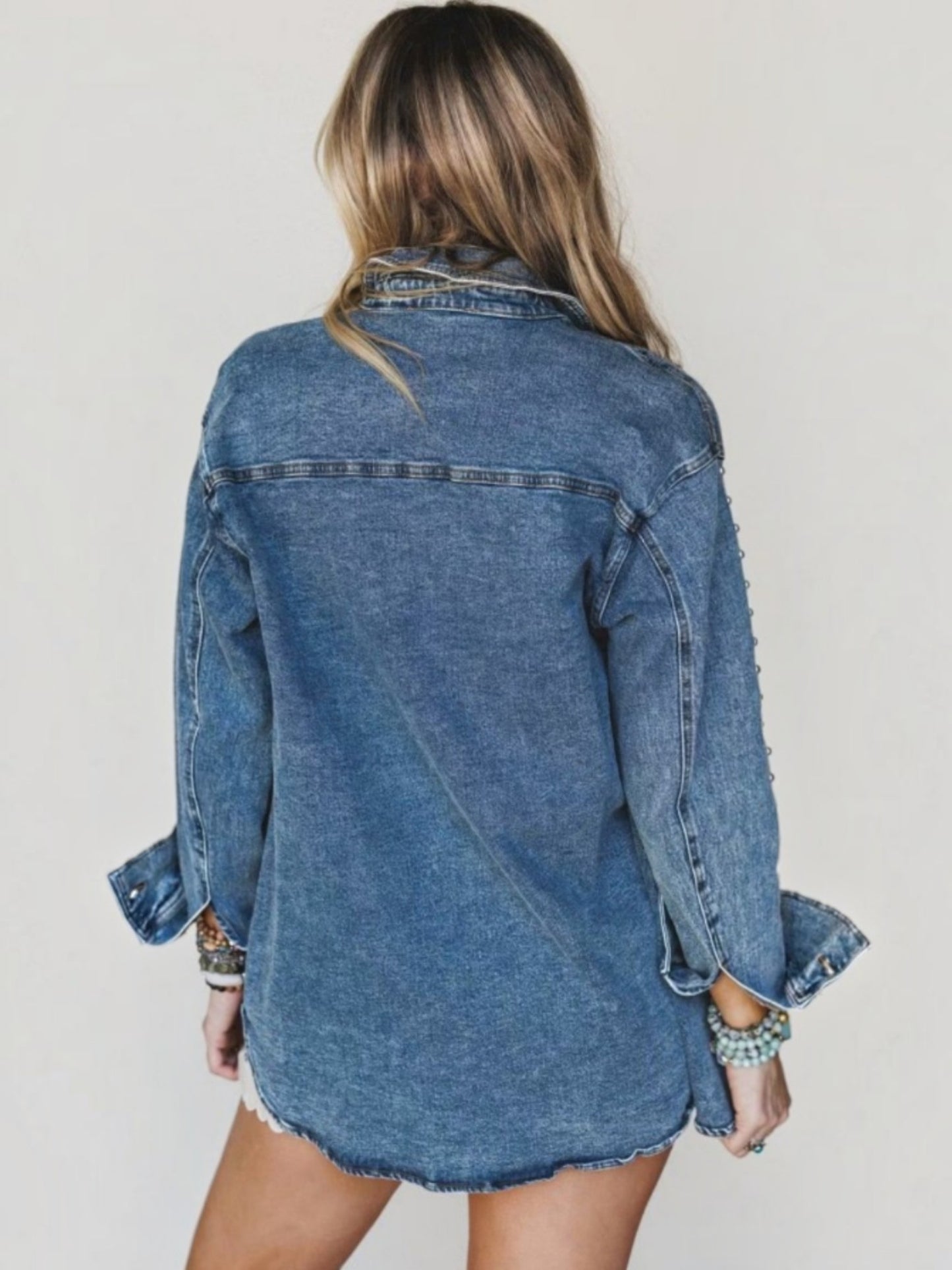 A woman shown from the back wearing an oversized blue denim shirt jacket with rolled cuffs and a raw hem, photographed against a plain light background.