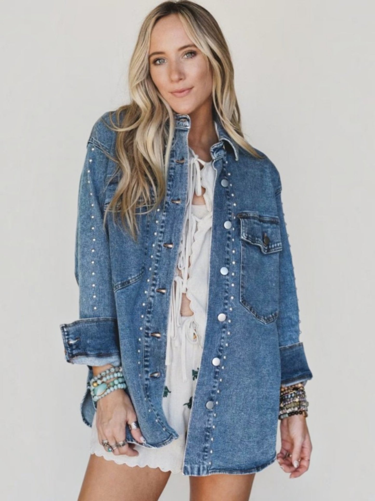 A woman modeling a blue denim jacket with silver studs, worn open over a white lace-up top and white shorts, against a plain light gray background.