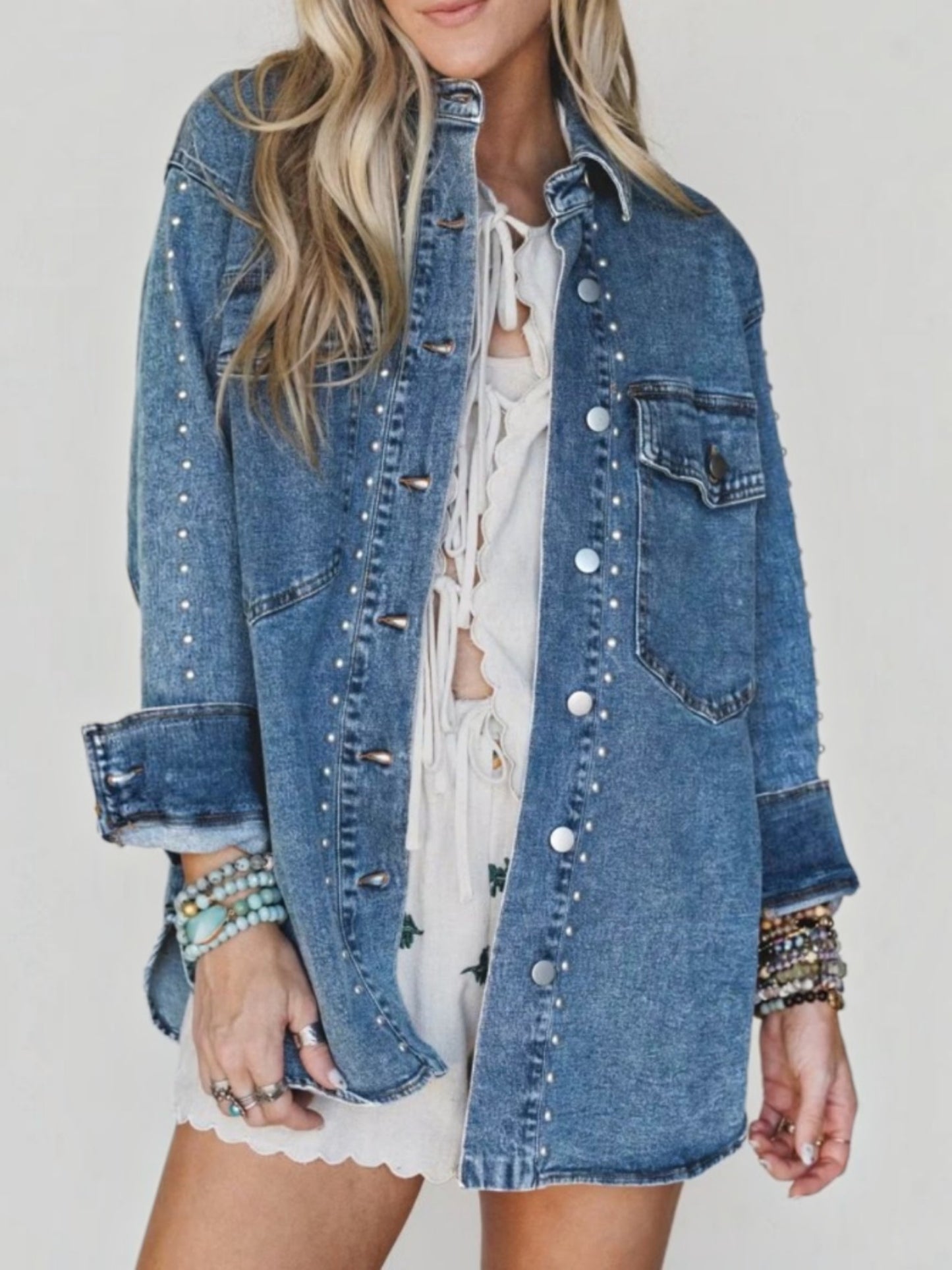 A woman wearing a blue denim jacket with silver studs and button closures over a white lace-up top and white skirt, photographed against a plain light background.