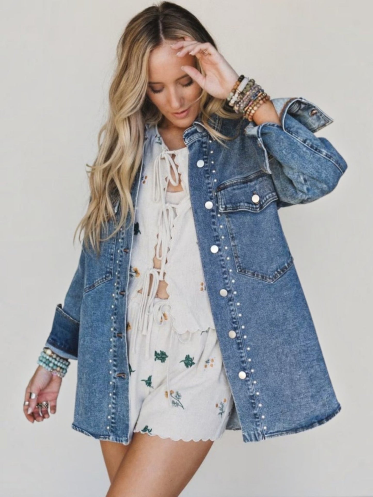 A woman wearing a light-wash blue denim jacket with silver studs and a white floral romper, posing against a light beige background.