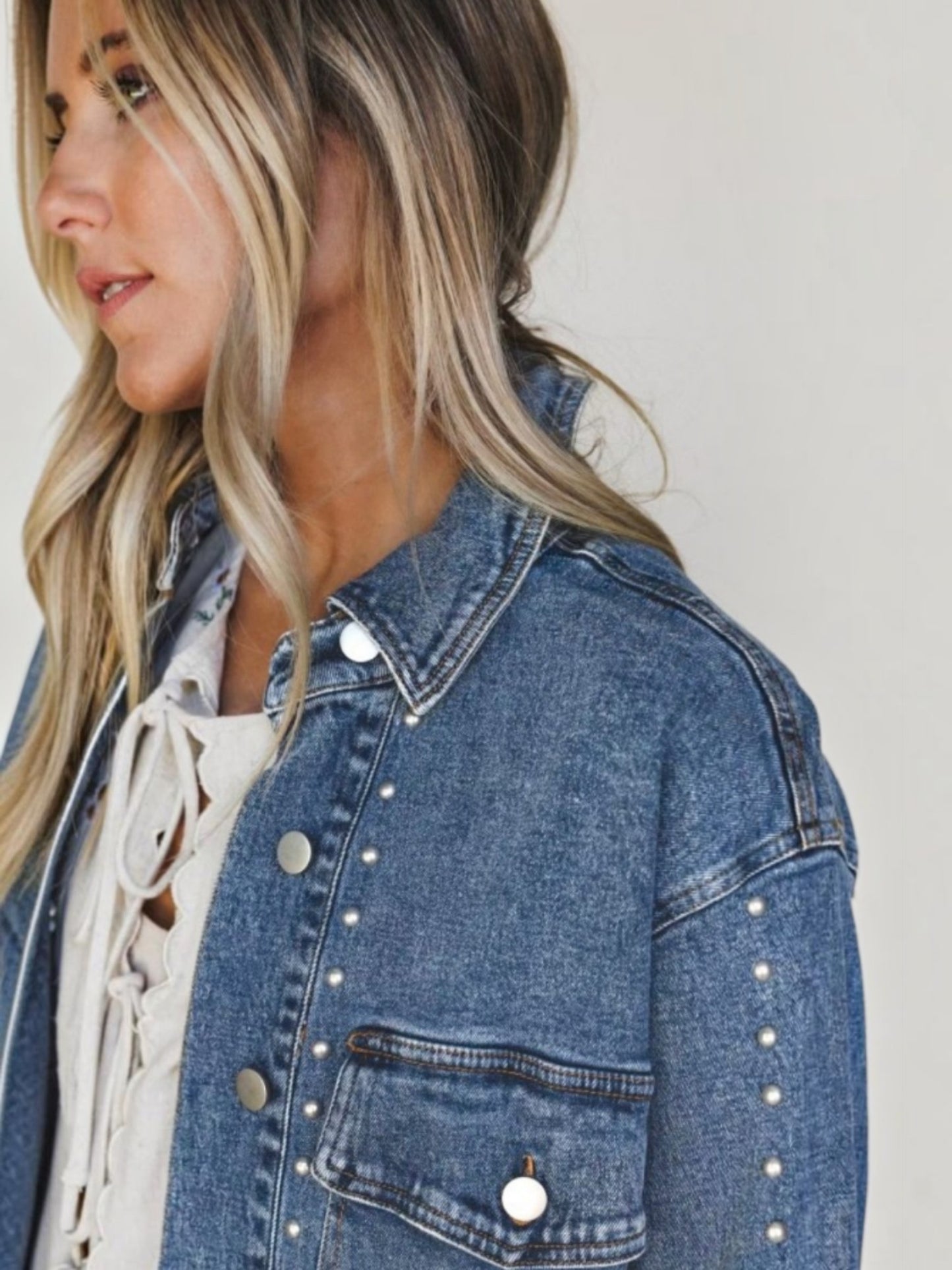 A close-up side view of a woman wearing a blue denim jacket with silver stud embellishments, layered over a white lace-up top, against a plain light background.