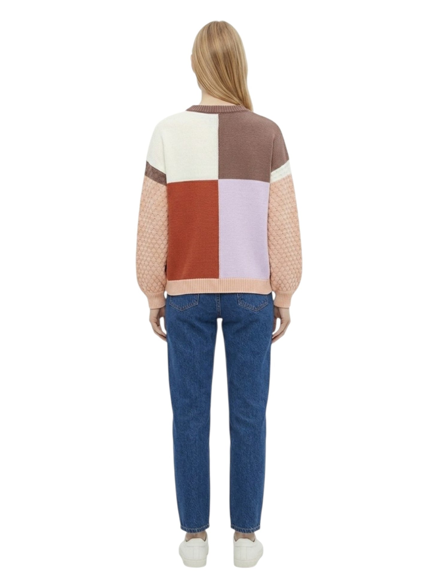 A woman viewed from the back wearing a color-block knit sweater with cream, brown, rust, pale lavender, and peach panels, paired with blue jeans and white sneakers on a white background.