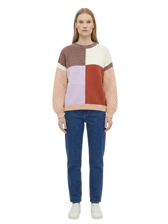 A woman stands in profile wearing a color-block knit sweater with brown, cream, peach, and light purple sections, blue jeans, and white sneakers against a plain white background front view.