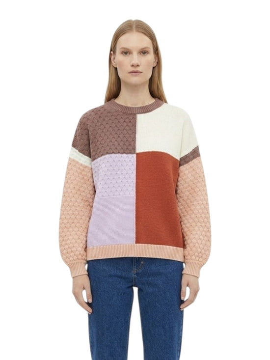 A woman wearing a color-block knit sweater with textured and smooth panels in brown, cream, light purple, rust orange, and peach, paired with blue jeans against a plain white background.