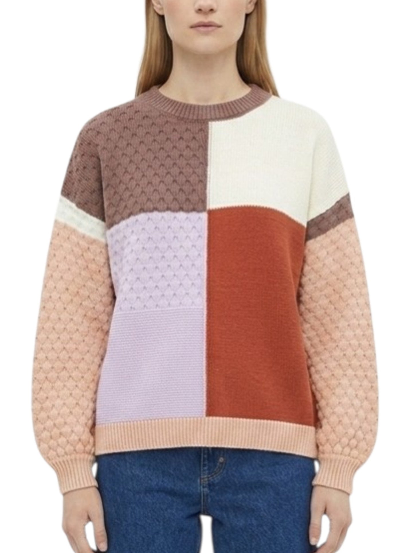 A woman wearing a color-block knit sweater with textured and smooth panels in brown, cream, light purple, rust orange, and peach, paired with blue jeans against a plain white background.