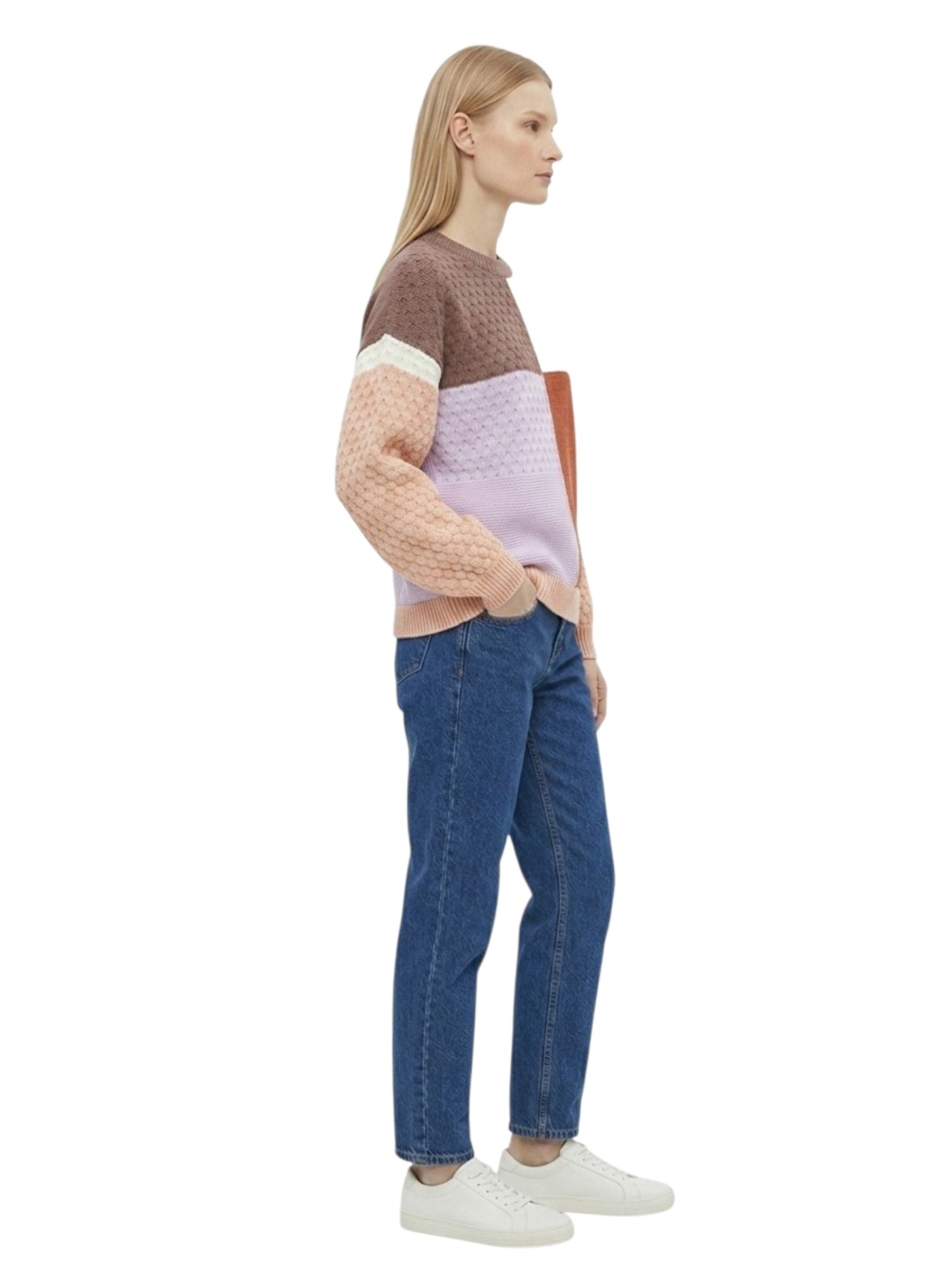 A woman stands in profile wearing a color-block knit sweater with brown, cream, peach, and light purple sections, blue jeans, and white sneakers against a plain white background.