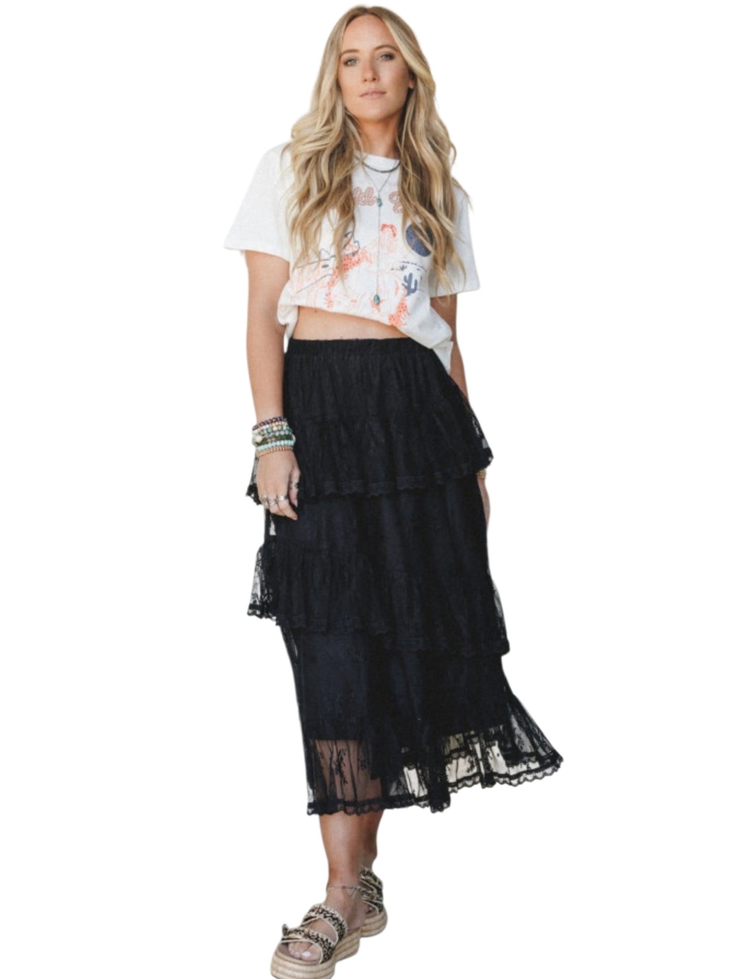 A woman modeling a black tiered tulle midi skirt with a ruffled hem, paired with a white graphic crop top and sandals, against a plain white background.
