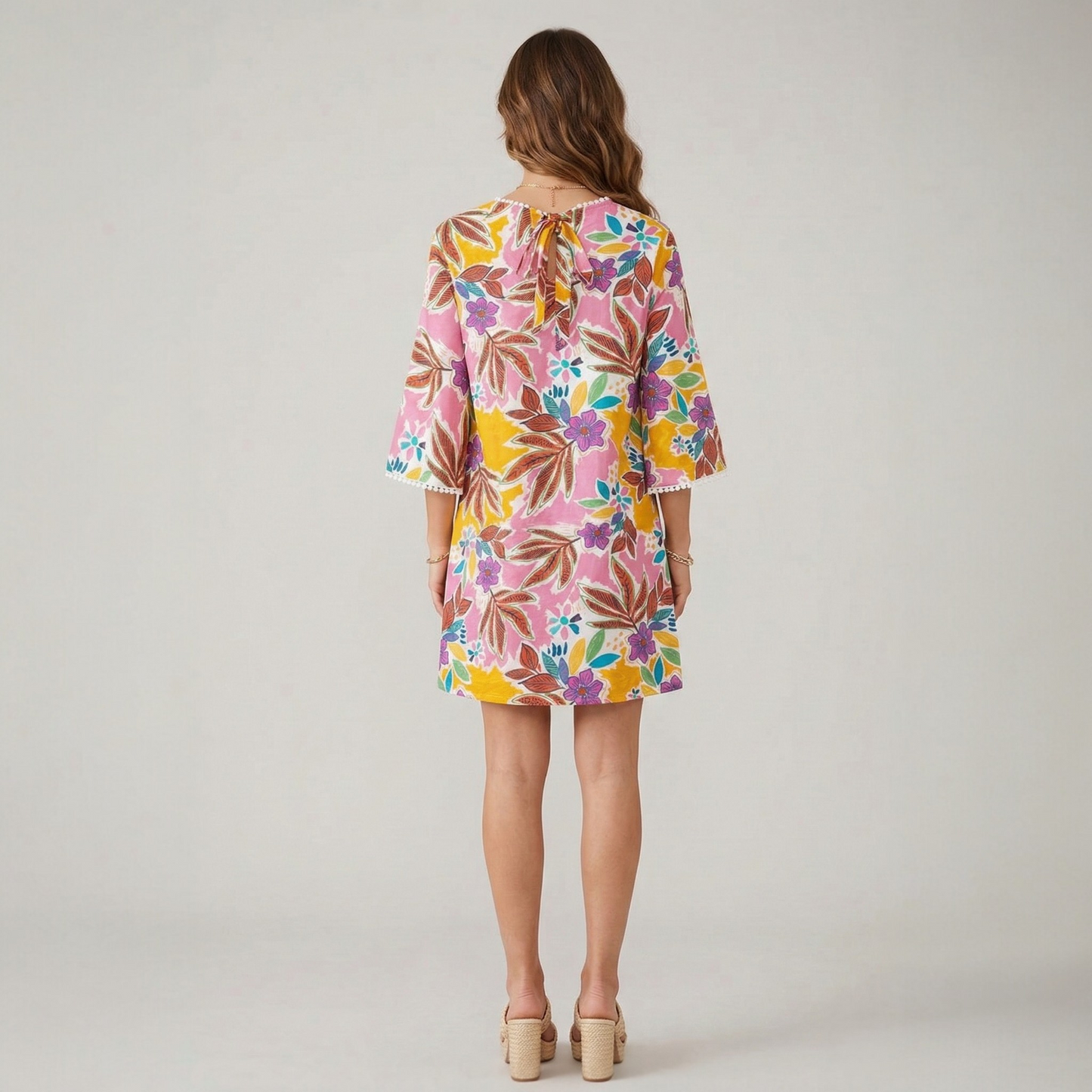 A model seen from the back wearing a floral print mini dress with pom-pom trim and a tie-neck detail, highlighting the flowy A-line silhouette.