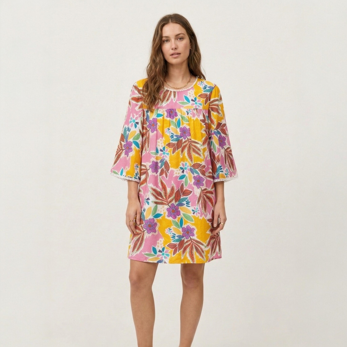 Full-length view of a model wearing a tropical floral mini dress with pom-pom trim, styled for a casual summer look.