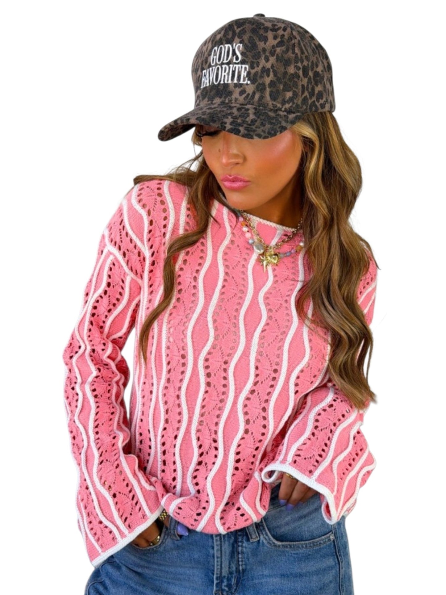 A woman wearing a pink and white wavy patterned long-sleeve top, blue jeans, and a brown leopard-print baseball cap with the text 'GOD'S FAVORITE' in white. The photo is shot against a solid white background.